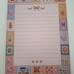 Può includere: Un foglio di carta da lettere rettangolare con un centro rosa chiaro a righe orizzontali e un bordo decorativo composto da vari riquadri. Il bordo presenta motivi ricamati di fiori, cuori, fiocchi e una piccola casa.