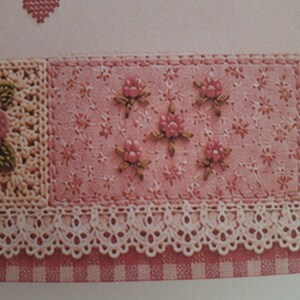 May include: A textile piece featuring a pink fabric panel with embroidered floral motifs, a cream-coloured crocheted lace border, and a pink and white checkered fabric edge. A small red cross-stitched heart is visible in the upper left corner.