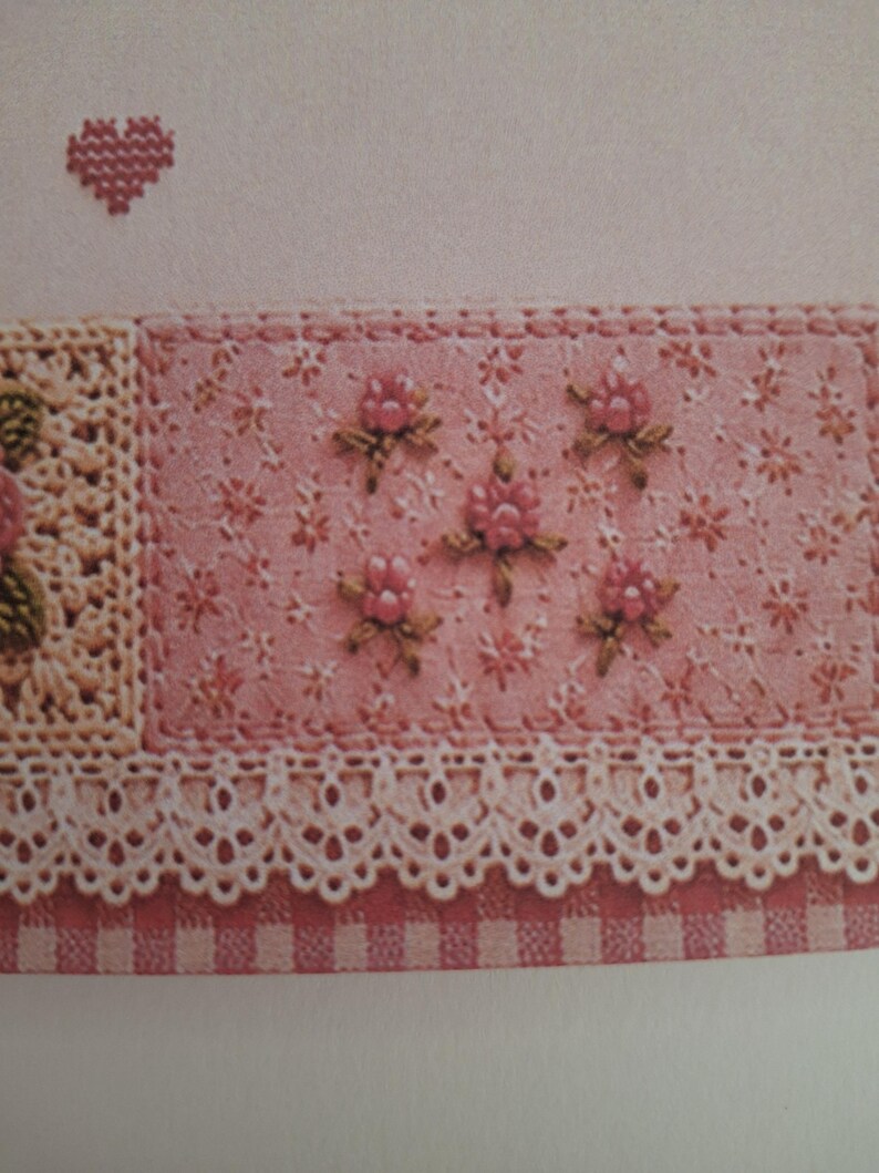 May include: A textile piece featuring a pink fabric panel with embroidered floral motifs, a cream-coloured crocheted lace border, and a pink and white checkered fabric edge. A small red cross-stitched heart is visible in the upper left corner.