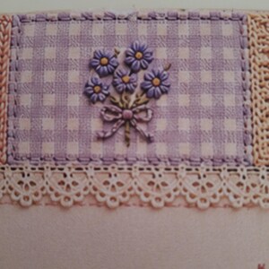 May include: A rectangular patch of light purple and white gingham fabric featuring an embroidered bouquet of purple flowers with yellow centres and a small bow. The patch is bordered by a cream-coloured lace trim and a textured, woven fabric edge.