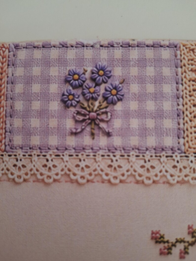 May include: A rectangular patch of light purple and white gingham fabric featuring an embroidered bouquet of purple flowers with yellow centres and a small bow. The patch is bordered by a cream-coloured lace trim and a textured, woven fabric edge.