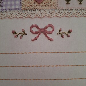 May include: A cross-stitch embroidery piece on light fabric featuring a central pink bow flanked by two small floral sprigs. Above, a white lace trim separates the lower section from a patterned border with a heart and checkered squares.