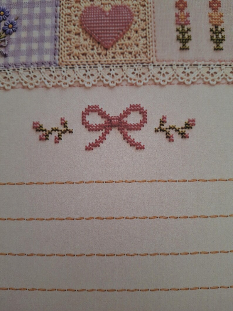 May include: A cross-stitch embroidery piece on light fabric featuring a central pink bow flanked by two small floral sprigs. Above, a white lace trim separates the lower section from a patterned border with a heart and checkered squares.