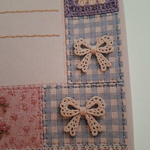 May include: A light pink stationery card featuring a patchwork design with blue and white gingham squares, a floral print section, and a crocheted flower motif. Two beige lace bows are attached to the gingham squares, and a lace trim borders the bottom.