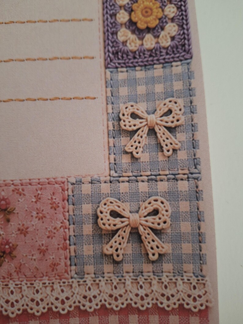 May include: A light pink stationery card featuring a patchwork design with blue and white gingham squares, a floral print section, and a crocheted flower motif. Two beige lace bows are attached to the gingham squares, and a lace trim borders the bottom.