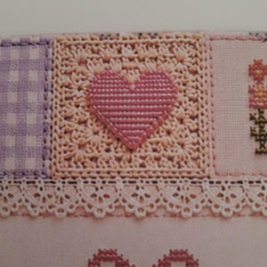 May include: A textile piece featuring a central square with a pink cross-stitched heart on a cream crocheted background, flanked by a purple gingham square and a floral motif. Below, a white lace trim sits above a pink bow and floral embroidery.