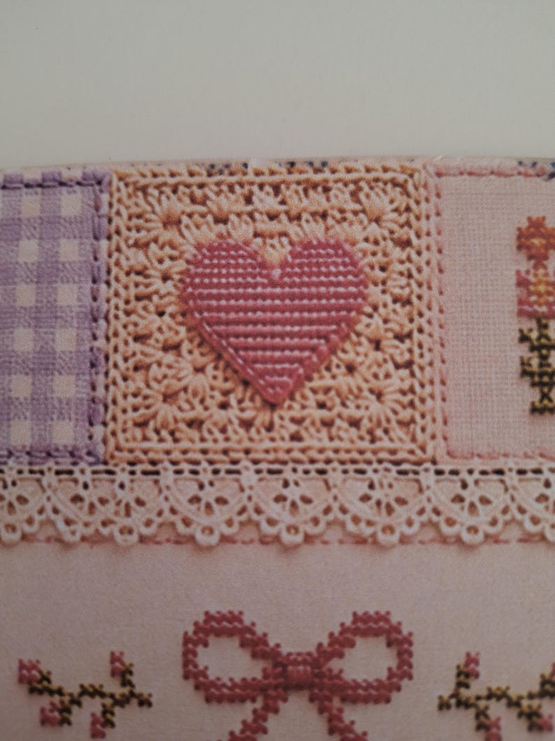 May include: A textile piece featuring a central square with a pink cross-stitched heart on a cream crocheted background, flanked by a purple gingham square and a floral motif. Below, a white lace trim sits above a pink bow and floral embroidery.