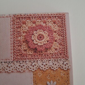 May include: A square crochet motif featuring a central pink and cream flower pattern, bordered by a delicate white lace trim. Below the lace, a mustard yellow panel displays a repeating white daisy print with additional white daisy embellishments.