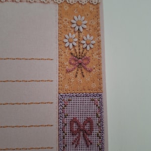 May include: A light pink stationery sheet featuring horizontal stitched lines, a white lace trim, and a vertical panel decorated with a bouquet of three white daisies tied with a pink bow, followed by a lavender cross-stitch square with a pink bow.