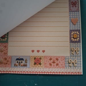 Puede incluir: Una hoja de papel de carta rayado con un borde decorativo de cuadrados estilo patchwork con patrones de punto de cruz de casas, flores, corazones y lazos. El papel tiene un fondo crema con tres peque&ntilde;os corazones rojos centrados abajo.