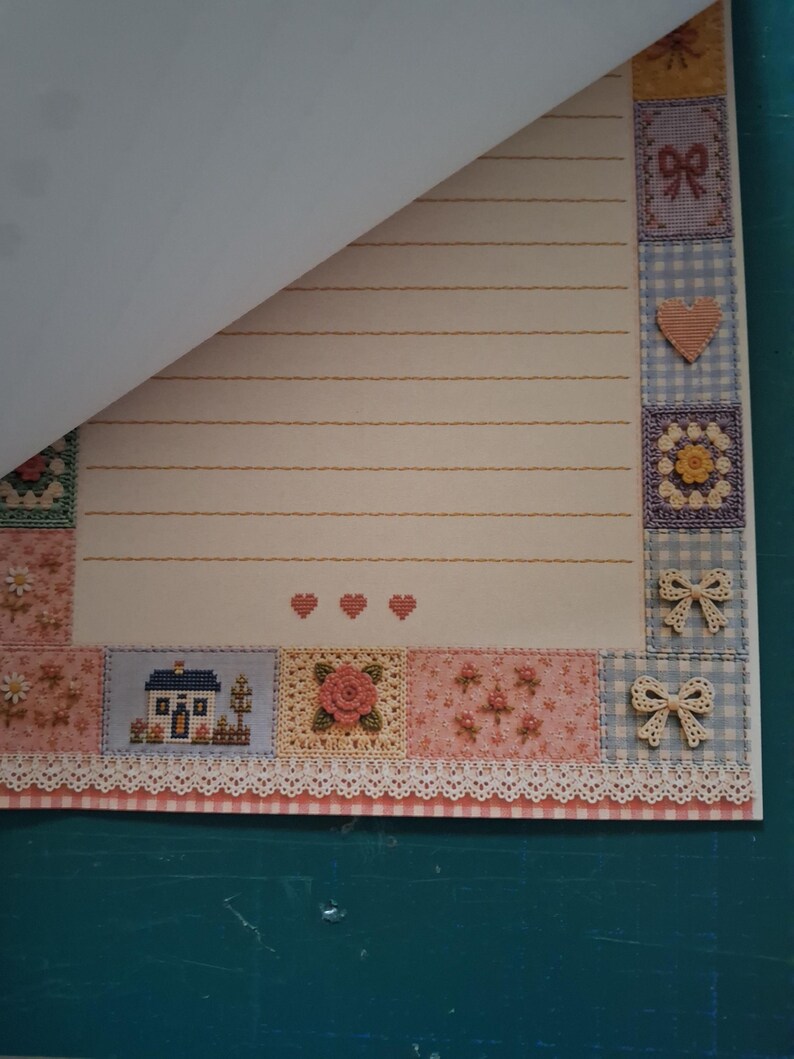Puede incluir: Una hoja de papel de carta rayado con un borde decorativo de cuadrados estilo patchwork con patrones de punto de cruz de casas, flores, corazones y lazos. El papel tiene un fondo crema con tres peque&ntilde;os corazones rojos centrados abajo.
