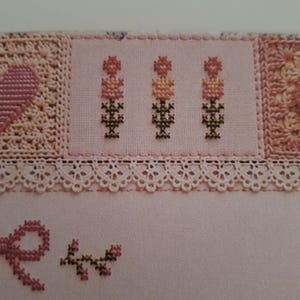 May include: A light pink fabric featuring cross-stitch embroidery of three small floral motifs in shades of pink, orange, and dark green. The design includes a delicate white lace trim border and a small pink stitched bow with a leafy branch.