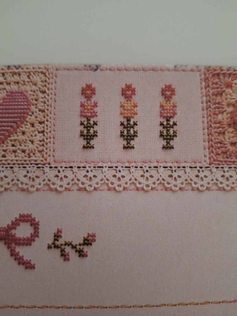 May include: A light pink fabric featuring cross-stitch embroidery of three small floral motifs in shades of pink, orange, and dark green. The design includes a delicate white lace trim border and a small pink stitched bow with a leafy branch.