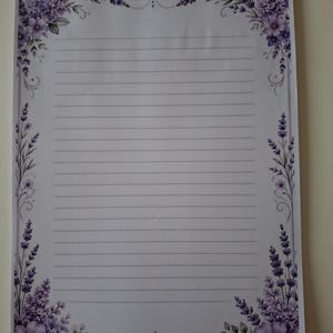 Op de afbeelding: Een rechthoekig vel wit briefpapier met horizontale lijnen om op te schrijven. De rand is versierd met paarse lavendelbloemen en groen blad, met symmetrische bloemstukken in elk van de vier hoeken van het papier.
