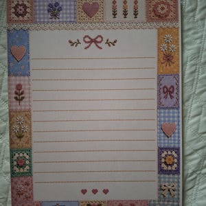 Puede incluir: Una hoja de papel de carta rectangular con un borde decorativo compuesto por varios cuadrados con patrones de punto de cruz, incluyendo flores, corazones, lazos y una casa. El centro tiene l&iacute;neas horizontales y un peque&ntilde;o icono de lazo.