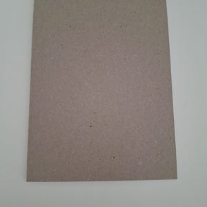 Puede incluir: Una hoja rectangular de cart&oacute;n de color marr&oacute;n claro con textura y un acabado mate plano. La superficie muestra sutiles variaciones de color y peque&ntilde;as motas oscuras, caracter&iacute;sticas del material de papel reciclado con bordes rectos.