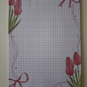 Può includere: Un foglio rettangolare di carta a quadretti con un bordo decorativo di tulipani rosa in stile acquerello negli angoli in alto a sinistra e in basso a destra, accentuato da nastri rosa fluenti e piccoli motivi a cuore su sfondo bianco.