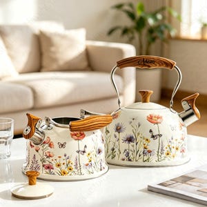 Könnte beinhalten: Zwei cremefarbene Teekessel mit einem bunten Wildblumen- und Schmetterlingsmuster. Die Kessel haben Griffe und Deckel in Holzoptik. Ein Kessel ist vollständig montiert, während der andere mit abgenommenem Deckel dargestellt ist.