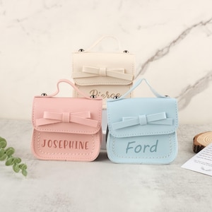 May include: Three small, pastel-colored purses with bow accents and top handles. The purses are in pink, light blue, and off-white. Each purse has a name printed on the front: "Josephine", "Ford", and "Pierce".