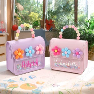 May include: Two small, pastel-coloured purses with flower embellishments and beaded handles. One is lavender with the name "Astrid", the other is pink with "Charlotte". Both have a clear front panel with glitter.