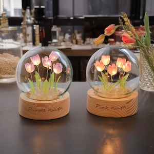 May include: Two glass domes with wooden bases, each containing a cluster of artificial tulips. The dome on the left has purple tulips and the base reads "Purple Tulips". The dome on the right has pink tulips and the base reads "Pink Tulips".