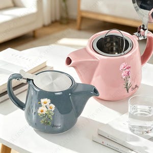 May include: A blue ceramic teapot with a white daisy floral print and the name Filippo, and a pink ceramic teapot with a pink carnation floral print and the name Sally. Both teapots feature a silver metal lid and a removable stainless steel infuser.
