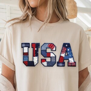 Op de afbeelding: Crèmekleurig T-shirt met het woord "USA" op de voorkant. De letters zijn gemaakt van patchworkstof in rood, wit en blauw patronen, inclusief sterren en strepen. Het shirt heeft korte mouwen.