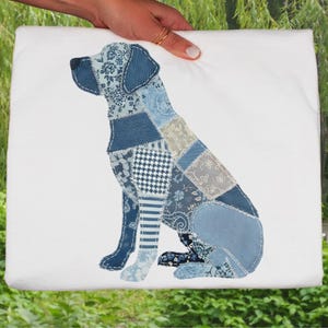 Op de afbeelding: Een witte draagtas met een patchwork hondenontwerp in verschillende blauwtinten. De hond zit, gemaakt van verschillende stoffen met patronen. De tas wordt vastgehouden door een hand met gouden ringen.