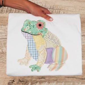 Peut inclure: Un carré de tissu blanc avec une application de grenouille colorée. La grenouille est faite de divers tissus à motifs, dont des motifs floraux, vichy et de la dentelle. L'œil de la grenouille est un bouton rose.