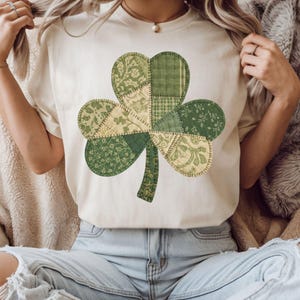 Può includere: T-shirt color crema con un grande design a trifoglio patchwork. Il trifoglio è composto da vari modelli di tessuto verde e crema, cuciti insieme. Un capo casual per l'uso quotidiano.