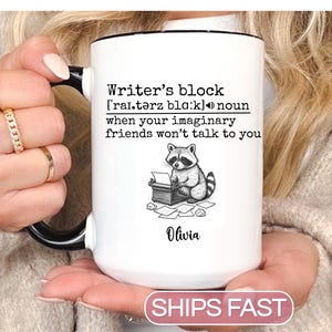 May include: White ceramic mug with a black handle and rim. The mug features the text "Writer's block" with a definition and a cartoon raccoon at a typewriter. The name "Olivia" is printed below the raccoon.