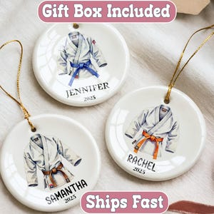 May include: Three white ceramic ornaments with gold hanging strings. Each ornament features a watercolor illustration of a karate uniform with a colored belt. Names and the year 2025 are printed below the uniform.