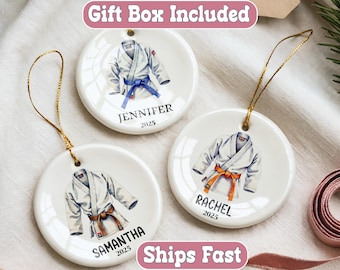May include: Three white ceramic ornaments with gold hanging strings. Each ornament features a watercolor illustration of a karate uniform with a colored belt. Names and the year 2025 are printed below the uniform.