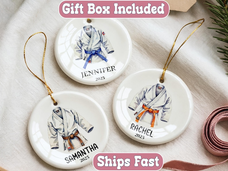 May include: Three white ceramic ornaments with gold hanging strings. Each ornament features a watercolor illustration of a karate uniform with a colored belt. Names and the year 2025 are printed below the uniform.