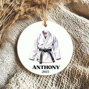 May include: A round, white ornament featuring a watercolor illustration of a white karate gi with a black belt. The name "ANTHONY" and the year "2025" are printed in black below the gi. A jute string is attached for hanging.