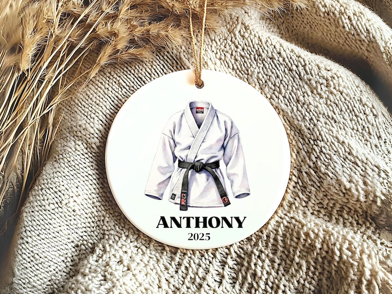 May include: A round, white ornament featuring a watercolor illustration of a white karate gi with a black belt. The name "ANTHONY" and the year "2025" are printed in black below the gi. A jute string is attached for hanging.
