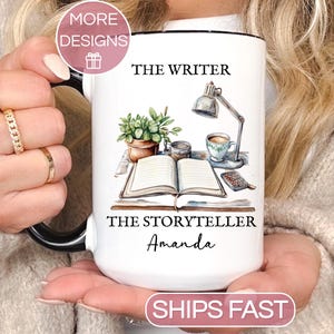 May include: White ceramic mug with black handle and rim, featuring a watercolor illustration of a writer's desk. The mug has the text "THE WRITER" and "THE STORYTELLER Amanda". A small pink circle with the words "MORE DESIGNS" is in the upper left corner.