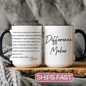 May include: White ceramic mug with a black handle and rim. The mug features the words "Difference Maker" in a handwritten font on one side and a longer inspirational quote on the other. The mug is displayed on a wooden surface.