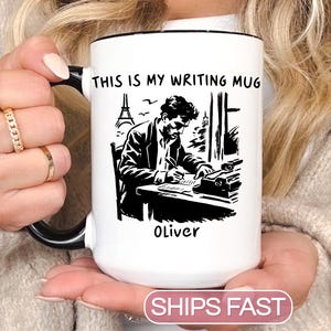 May include: White ceramic mug with a black handle and rim. The mug features a black and white illustration of a writer at a desk with a typewriter and the text "THIS IS MY WRITING MUG" and the name "Oliver".