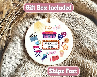 May include: A white, round ornament with theater-themed illustrations, including a clapperboard, stage, masks, and popcorn. The name "Melanie" and the year "2025" are displayed. The ornament has a string for hanging and the text "Gift Box Included".