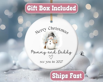 May include: A round white ceramic Christmas ornament hanging from a gold ribbon. The center features a watercolor snowman wearing a gray hat and holding a broom. Text reads Merry Christmas, Mommy and Daddy, a heart symbol, and see you in 2027.