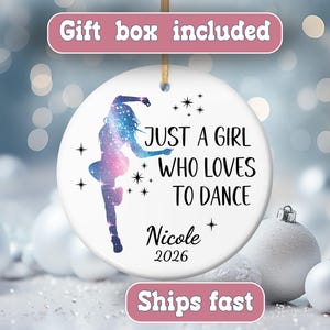 May include: A round white ceramic ornament featuring a galaxy-patterned silhouette of a dancer, black text reading "JUST A GIRL WHO LOVES TO DANCE", the name "Nicole", and the year "2026", accented with small black star icons.