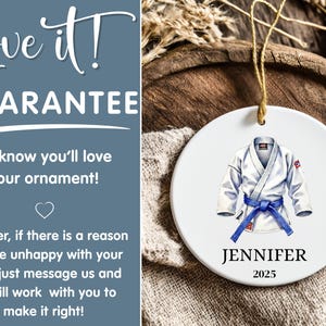 May include: A white ceramic ornament featuring a watercolor illustration of a judo gi with a blue belt. The ornament includes the name "JENNIFER" and the year "2025". It hangs by a gold string.