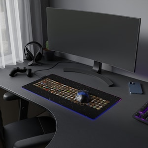 May include: A black gaming mouse pad with a colorful design of many small icons. A blue and black gaming mouse is on the mouse pad. A black computer monitor, a black headset, and a black smartphone are also on the desk.