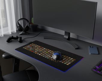 May include: A black gaming mouse pad with a colorful design of many small icons. A blue and black gaming mouse is on the mouse pad. A black computer monitor, a black headset, and a black smartphone are also on the desk.