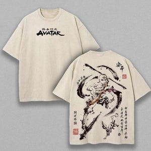 以下が含まれることがあります： ベージュ色のTシャツ。前面には黒字で「AVATAR」の文字と日本語が書かれています。背面には、棒を持った人物の黒と茶色のイラスト、日本語のテキスト、赤いスタンプが描かれています。