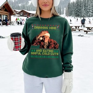 Puede incluir: Una sudadera verde oscuro con el texto "DRINKING WINE?" y "AND EATING SINFUL COLD CUTS" en blanco y rojo. La sudadera presenta un gráfico de un hombre con un gorro de Papá Noel. La persona lleva guantes blancos y sostiene una taza roja y negra.