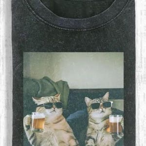 以下が含まれることがあります： サングラスをかけた2匹の猫がビールジョッキを持っているグラフィックが特徴の濃いグレーのTシャツ。猫は薄茶色と灰色のトラ柄です。画像はヴィンテージで、少し色あせた外観です。