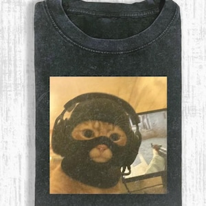 May include: Dark gray t-shirt featuring a cat wearing a black balaclava and headphones. The cat is in front of a computer screen. The t-shirt has a crew neck and short sleeves. The image is printed on the front of the shirt.