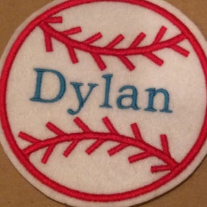 Puede incluir: Béisbol de fieltro blanco con costuras rojas y el nombre "Dylan" bordado en hilo azul.